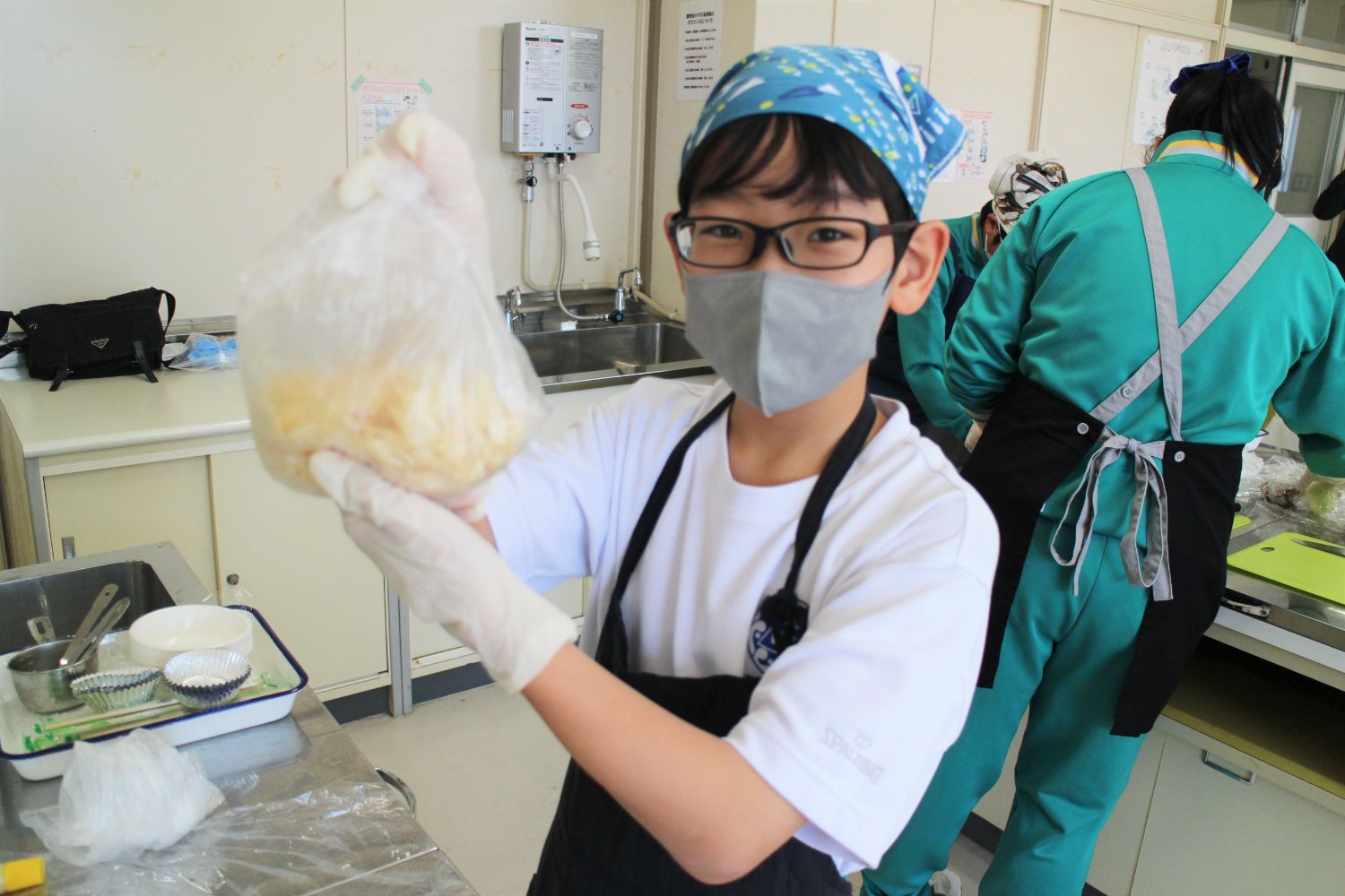 マスクとエプロンをつけた子どもが、ビニール袋に入った衣を見せながら笑顔を見せている写真