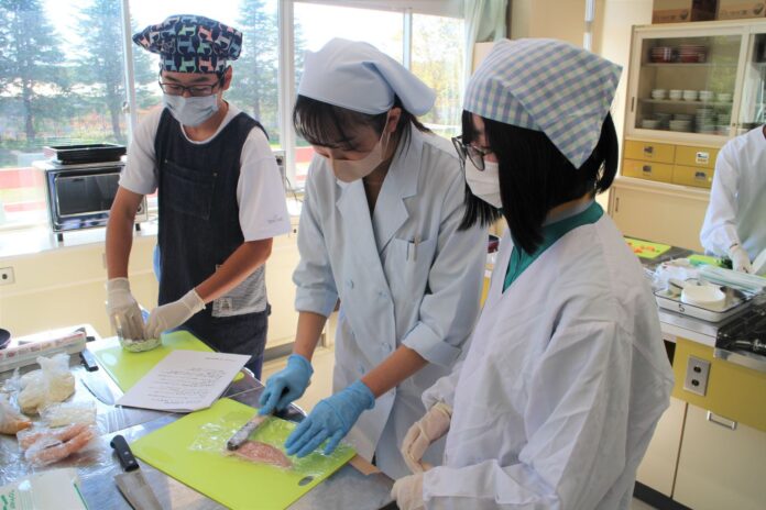 調理実習室で三角巾とマスクを着けた生徒たちが、白衣姿の生徒と共に食材を下ごしらえしている写真