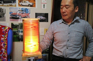 木材を透かして柔らかく発光している縦型の円筒形構造の手作りの木製ランプを渡部正行さんが持っている写真