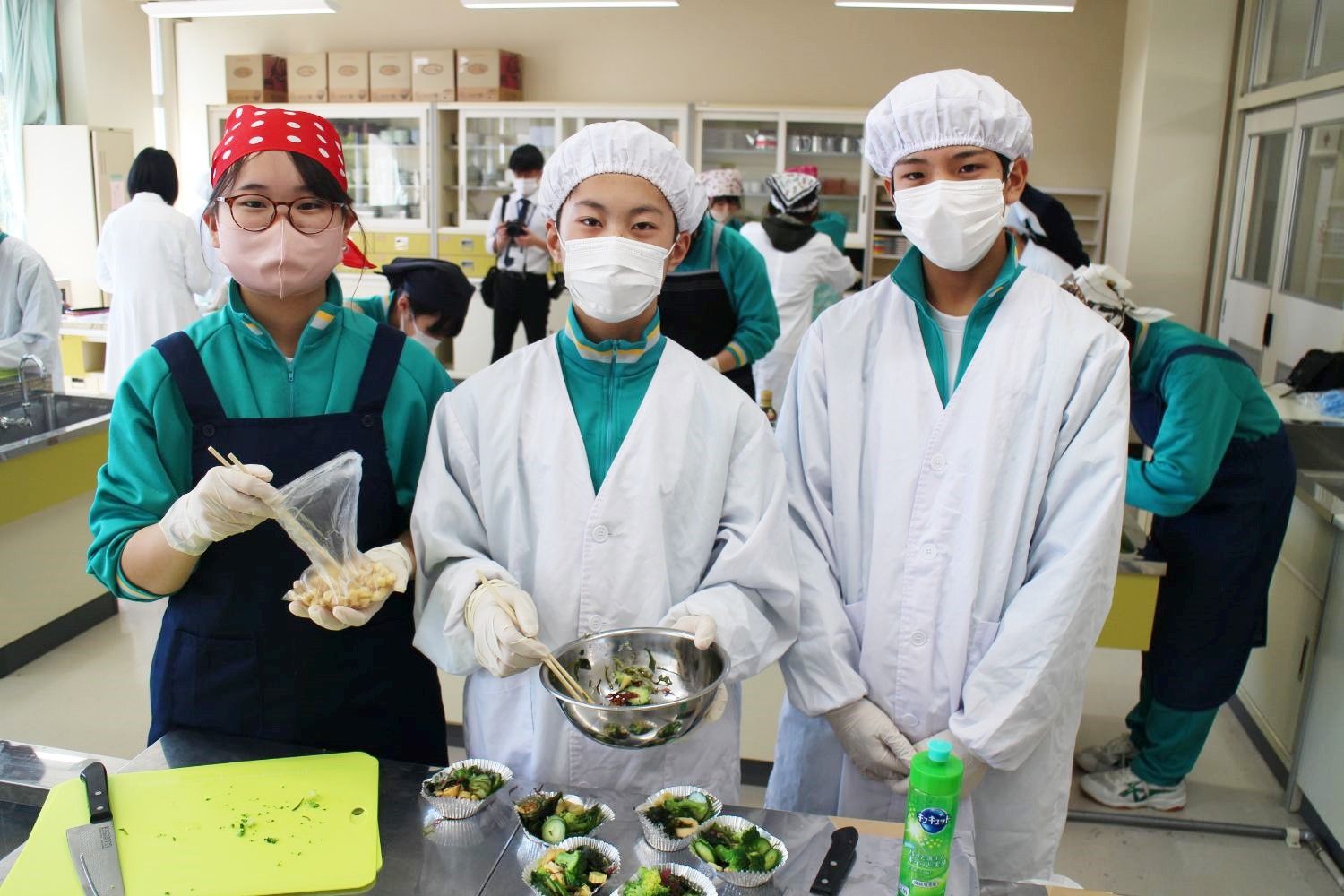 3人の中学生が手袋とマスクを着用し、中央の生徒がサラダを入れたステンレスボウルを持ち、左の生徒がクルトンが入った袋を持っている様子の写真