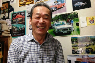 ラシックカーの写真やポスターの前でチェック柄のシャツを着た笑顔の渡部正行さんの写真