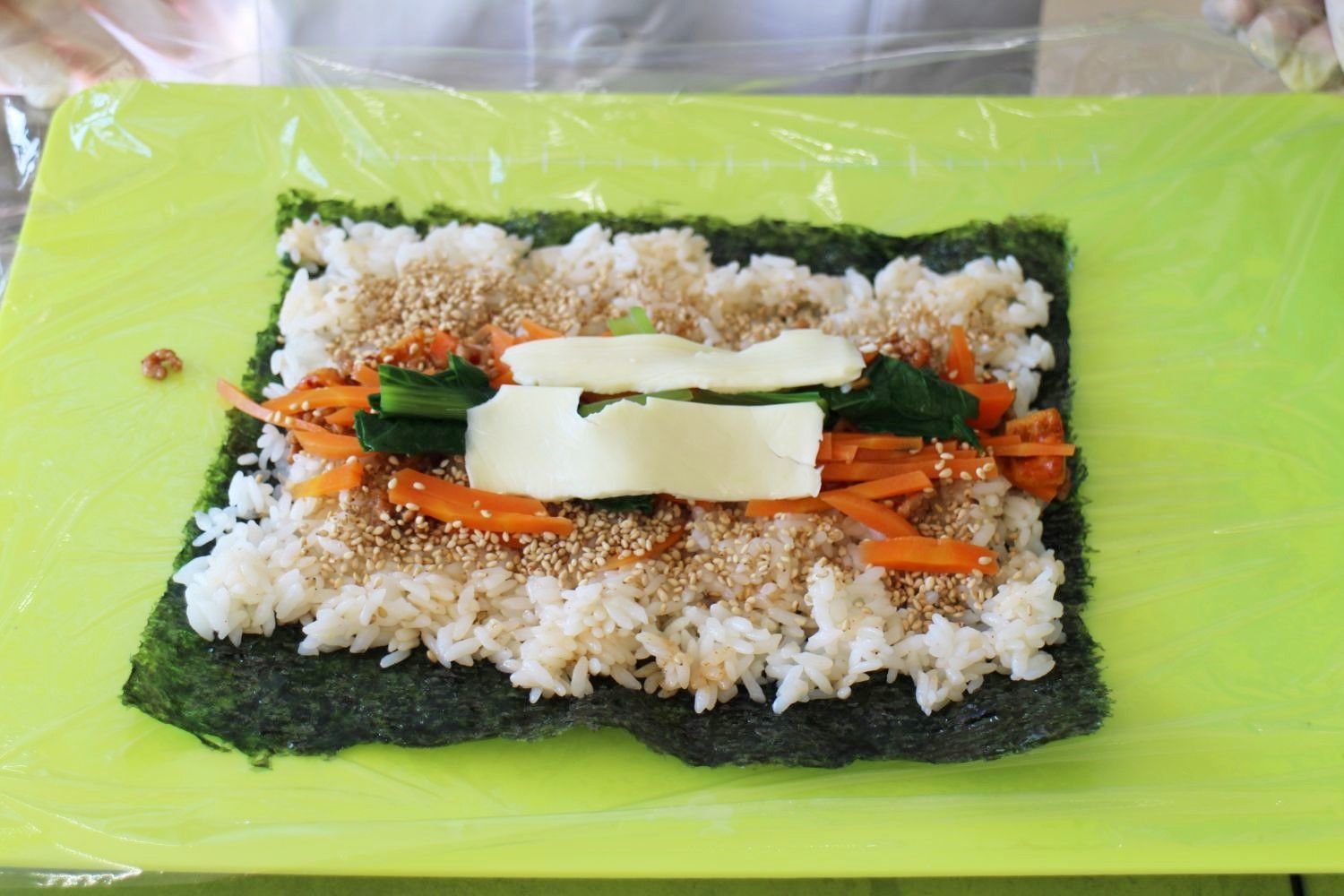 海苔の上にご飯を広げ、その上にニンジンやホウレンソウ、チーズ、白ごまをのせて巻く準備をしている様子の写真