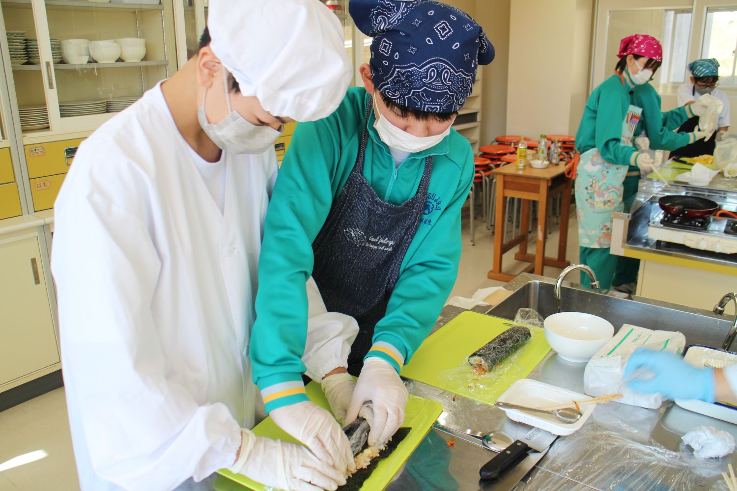 手袋とマスクを着けた2人の子どもが協力して巻き寿司を巻いている様子の写真