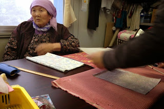 机の上に布を広げて作業する人と、その様子を見守る高齢の女性がいる写真