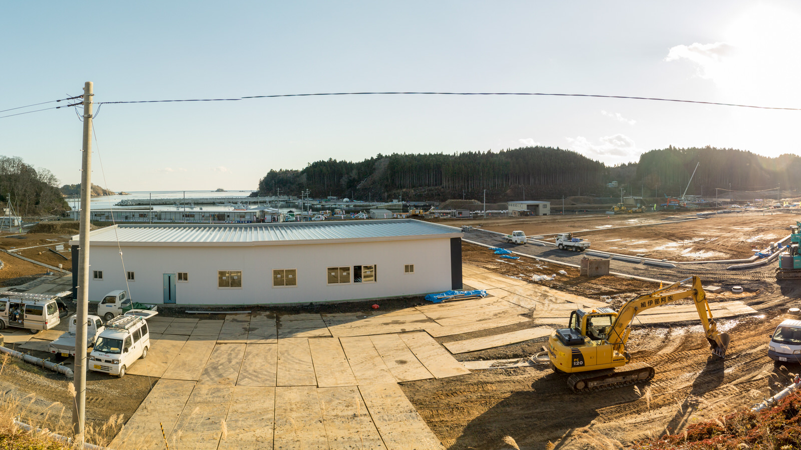 手前には白い建物が建って周囲にはトラックやショベルカーなどの車両が複数あり、現場で作業が行われている様子が見られ、背景にはさらに建設活動が広がっており、森林地帯と晴れた空が遠くに見える写真