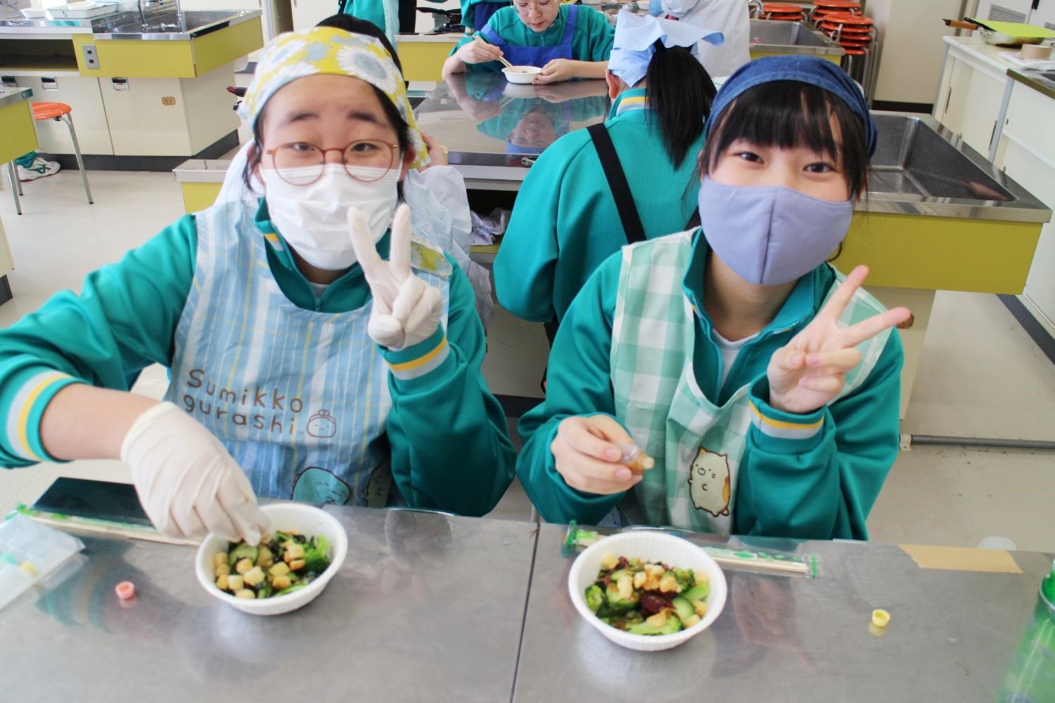 エプロン姿の女子生徒2人が、机に並べられたサラダ入りの器を前にして、ピースサインをしながら笑顔でポーズをとっている写真