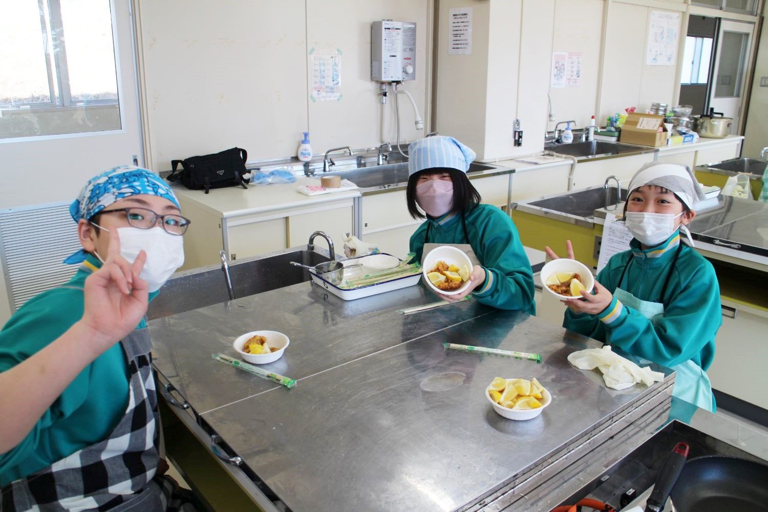 調理実習室のテーブルで、揚げ物の入った器を手に持って見せながら、3人の生徒が笑顔でピースサインをしている写真