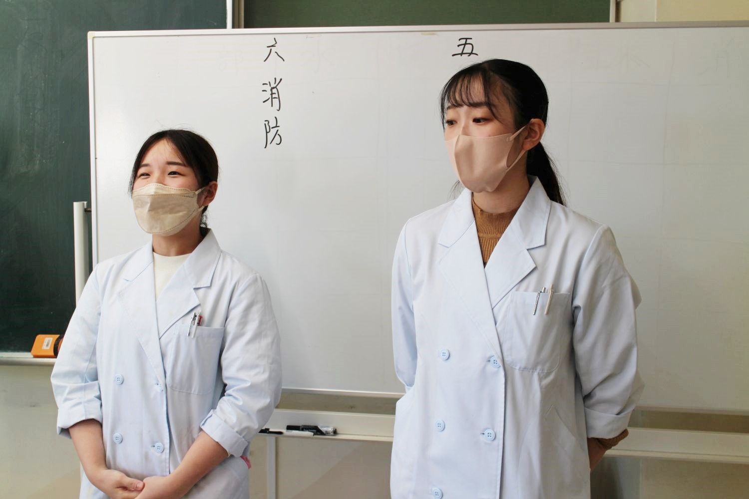 白衣姿でマスクを着けた女子学生2人が教室前方に立ち、発表をしている写真