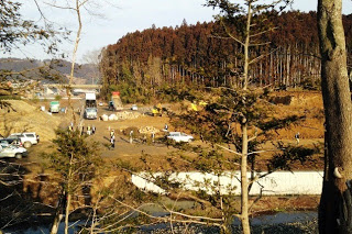 木々の間から、造成工事中の現場にトラックなど複数の車両が停まっており、周囲には作業をしている人の姿がある様子を見下ろした写真