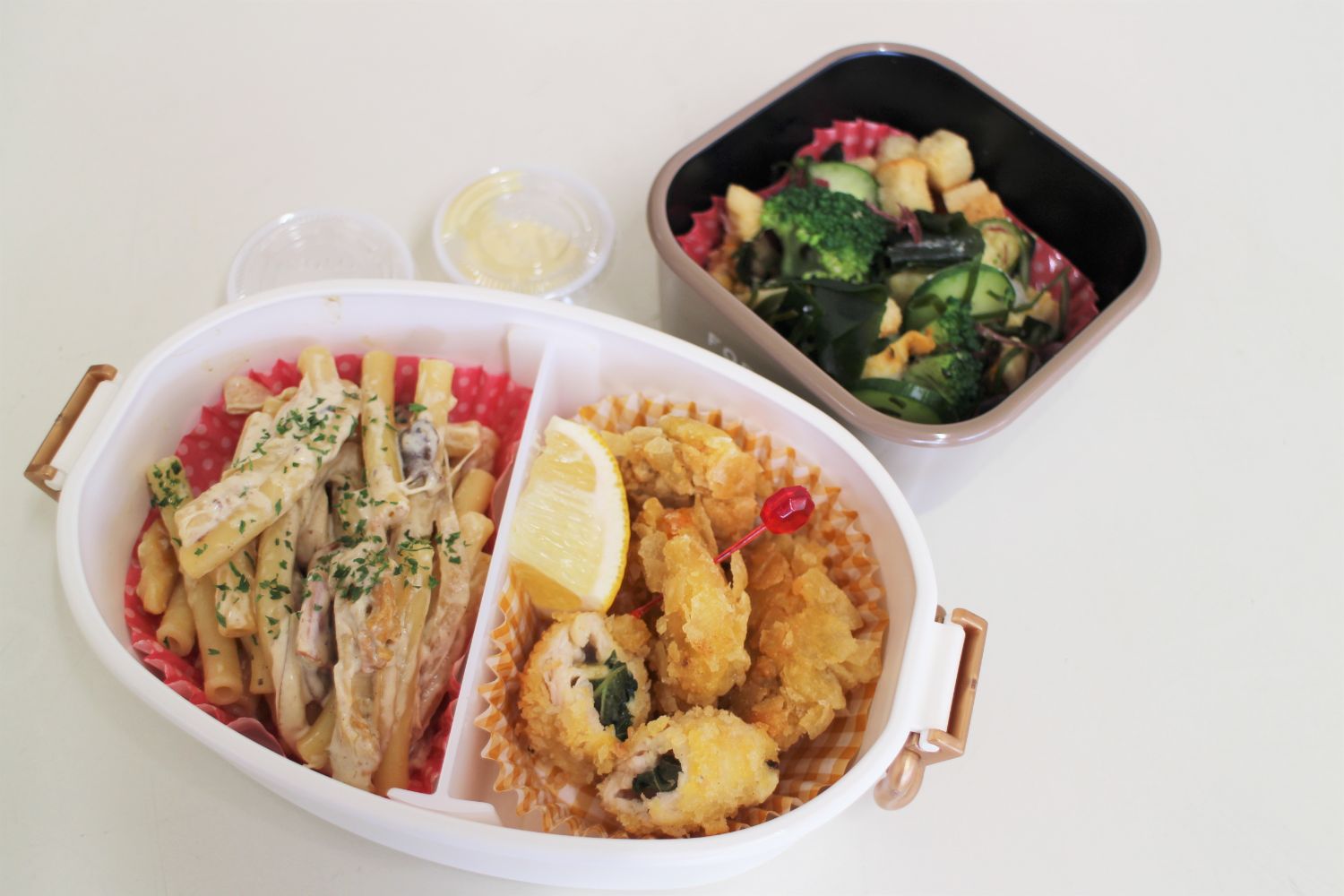 マカロニのクリームパスタ、ホヤの唐揚げ、しそ巻きホヤフライ、レモンが仕切られて入っている弁当と、サラダが入った小さな容器が並んでいる写真