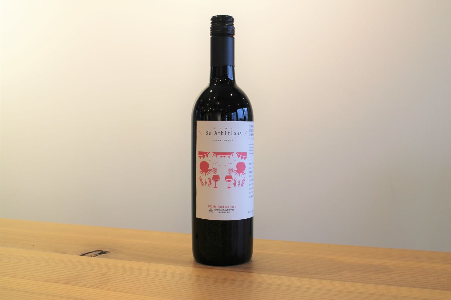 「Be Ambitious」と書かれ、ピンク色でイラストが描かれた白いラベルが貼られた黒いワインボトルが1本テーブルの上に立っている写真