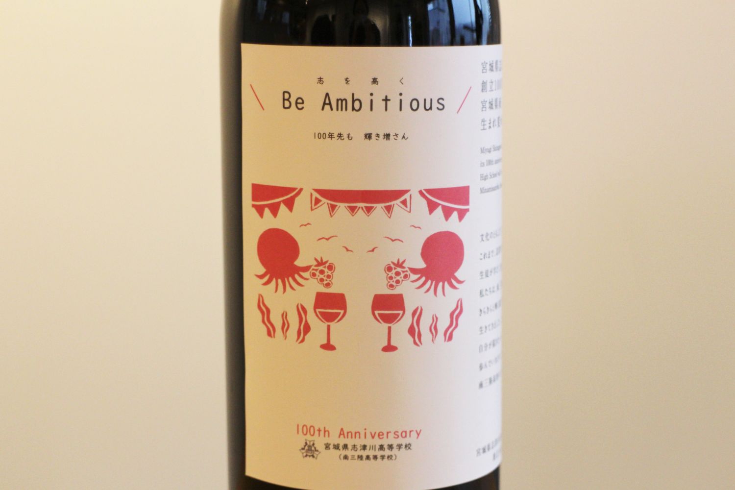 「Be Ambitious」と書かれたラベルの拡大で、タコやワイングラス、海藻などのイラストがピンク色で描かれている、志津川高校の生徒がデザインした記念ワインラベルの写真