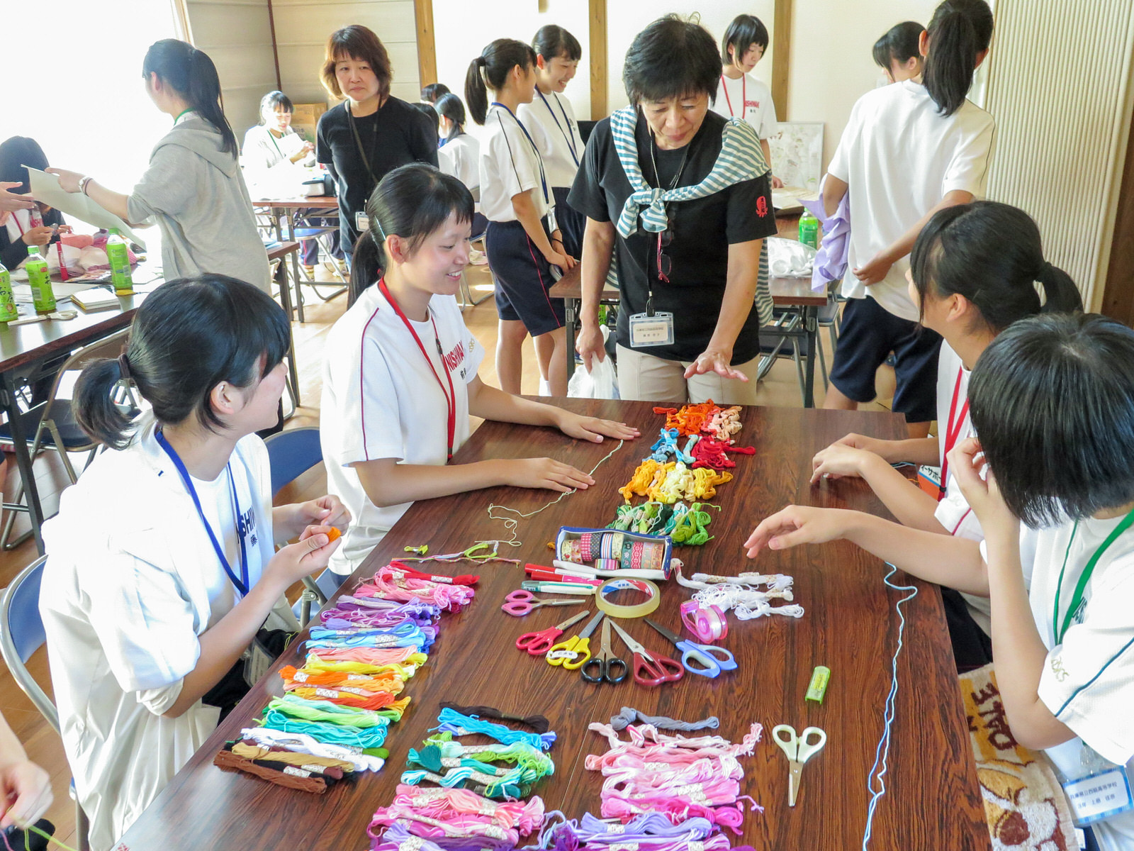 明るい室内で、机いっぱいに広げられたカラフルな刺しゅう糸や道具を囲んで女子高校生たちと指導の女性たちが楽しそうに会話しながら手芸をしている写真