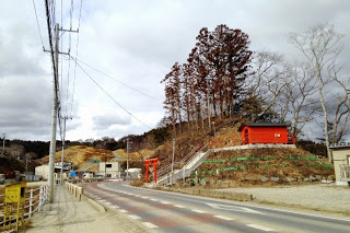 舗装道路のカーブの手前右側に赤い鳥居があり、奥の木々が生えた小高い丘の中腹に赤い本殿の小森御前社がある様子の写真