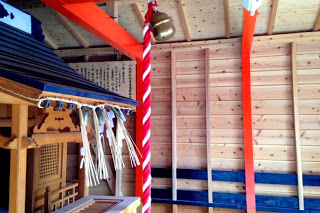 天井からは赤と白のねじれた縄が吊るされており、上部には鈴が取り付けられていて左側には紙垂が垂れ下がっている祭壇がある本殿の中を写した写真