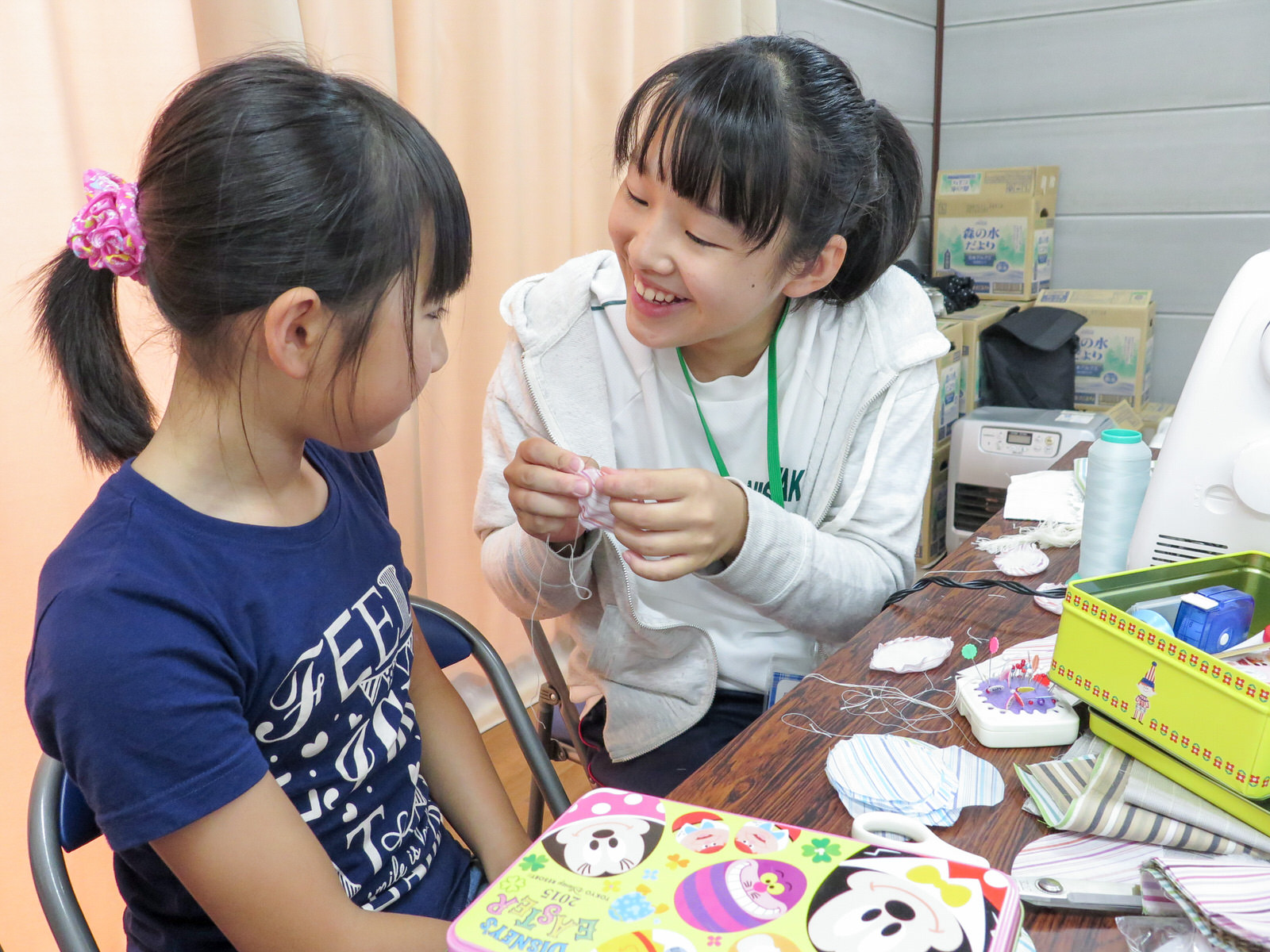 道具が置かれたテーブルで隣に座った小学生の女の子に笑顔で話しかけながら手芸をしている女子高校生の写真