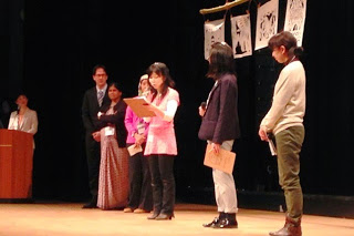 ステージ上に6人の人物が立っており、左側には演台があり背景には、絵や文字が描かれた3枚の垂れ幕が掛けられている写真