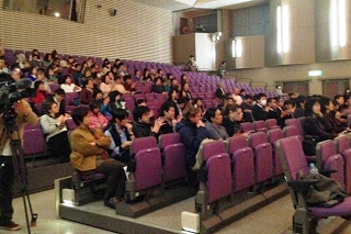 白い壁と照明のある講堂の内部で、多くの人々が紫色の椅子に座っている様子を写した写真