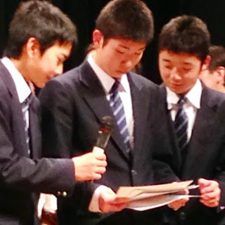 3名の男子学生の一番左の人がマイクを持ち、中央の学生がたくさんの用紙を持ちながら3人で覗き込んでいる写真