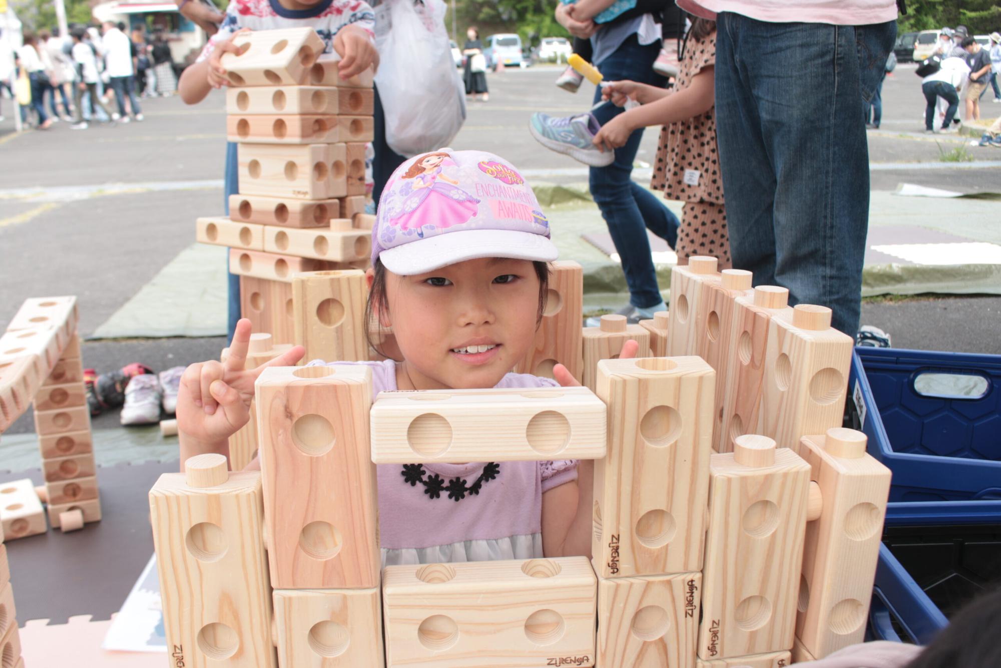 帽子をかぶった女の子が、高く積み上げた木のブロック越しにピースサインをしている写真