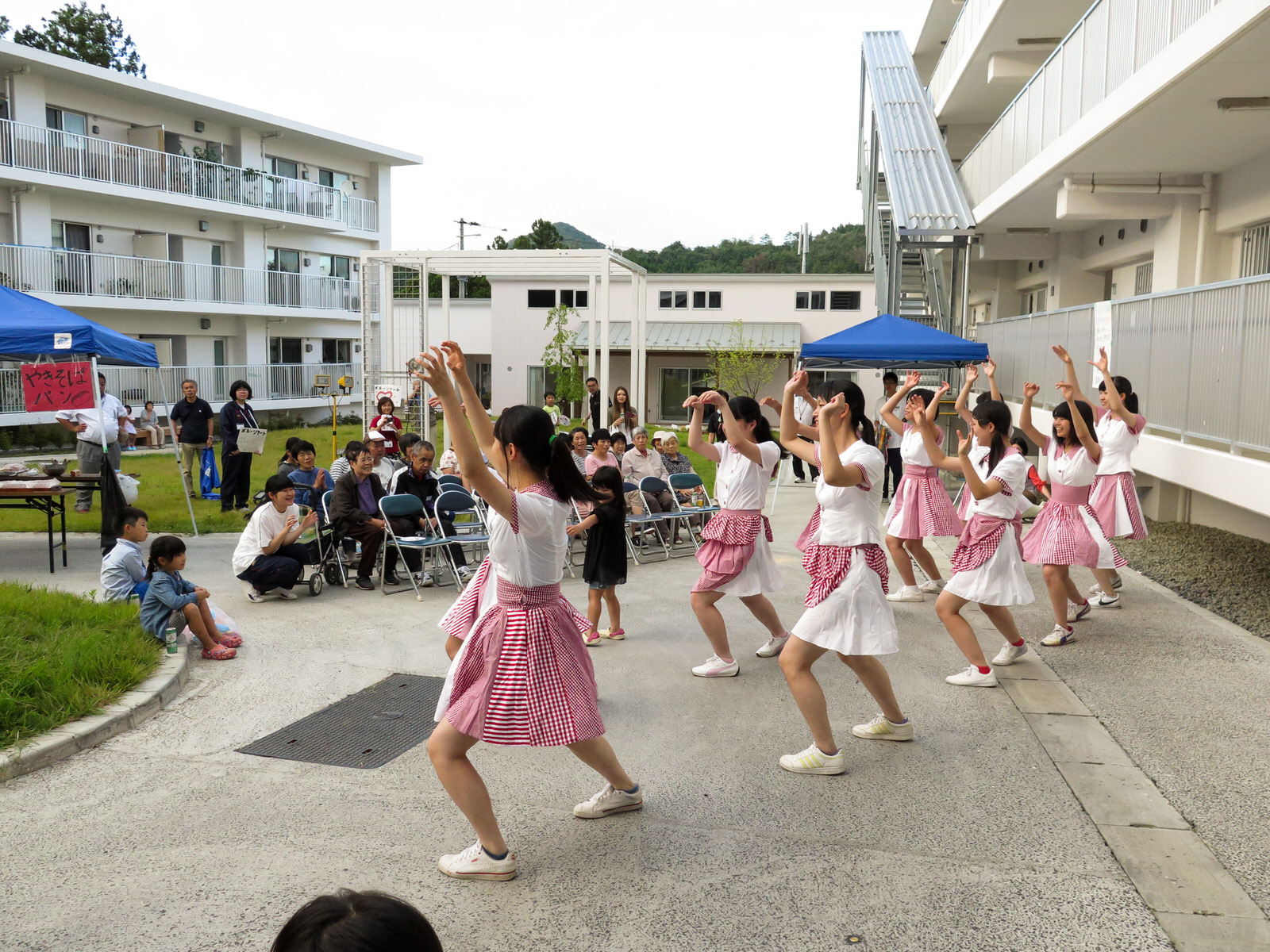 災害公営住宅の中庭で白いシャツと赤いチェック柄のスカートを着た女子生徒たちがダンスを披露し、それを椅子に座った高齢者や子どもたちが楽しそうに見ている写真