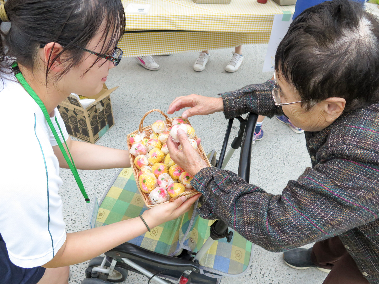 女子高校生が差し出したかごから、歩行器を押した高齢の女性いくつかのカラフルな手毬寿司を受け取っている写真