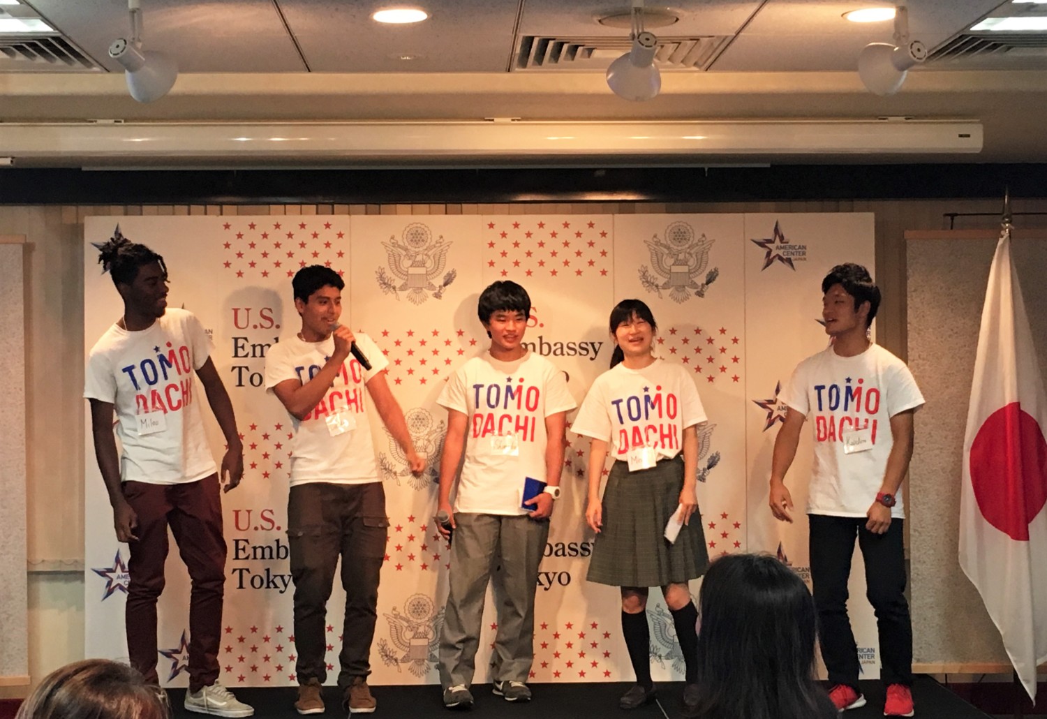「TOMODACHI」と書かれたお揃いのTシャツを着た5人の若者がバックパネルの前に一列に並び、左から2番目の男性がマイクを持ちプレゼンテーションを行っている様子の写真