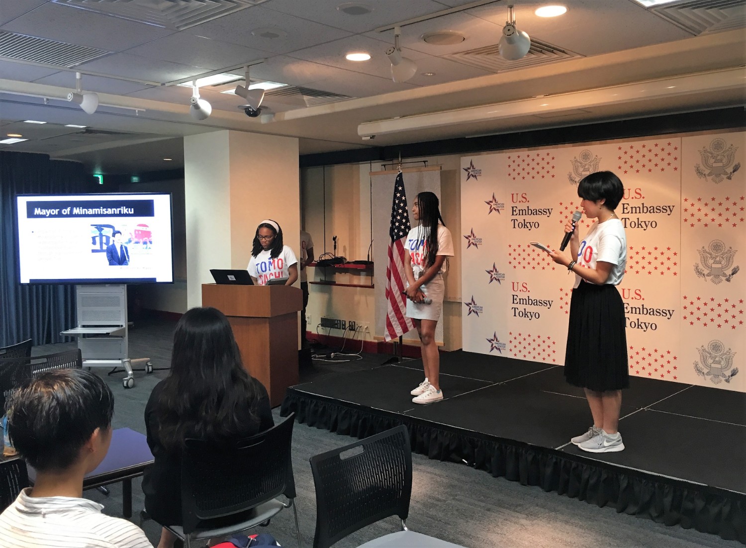 資料が映し出されたスクリーンの傍にあるステージで、「TOMODACHI」と書かれたTシャツを着た3人の若者が発表をしている写真