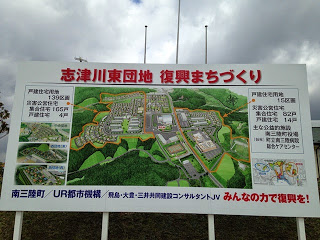 団地全体の完成予定図と説明が書かれている、「志津川東団地 復興まちづくり」と題されたカラフルな案内板の写真