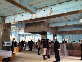 数人の人々がヘルメットや安全装備を着用しており、天井や梁には青い断熱材が覆われ内部見学をしている写真