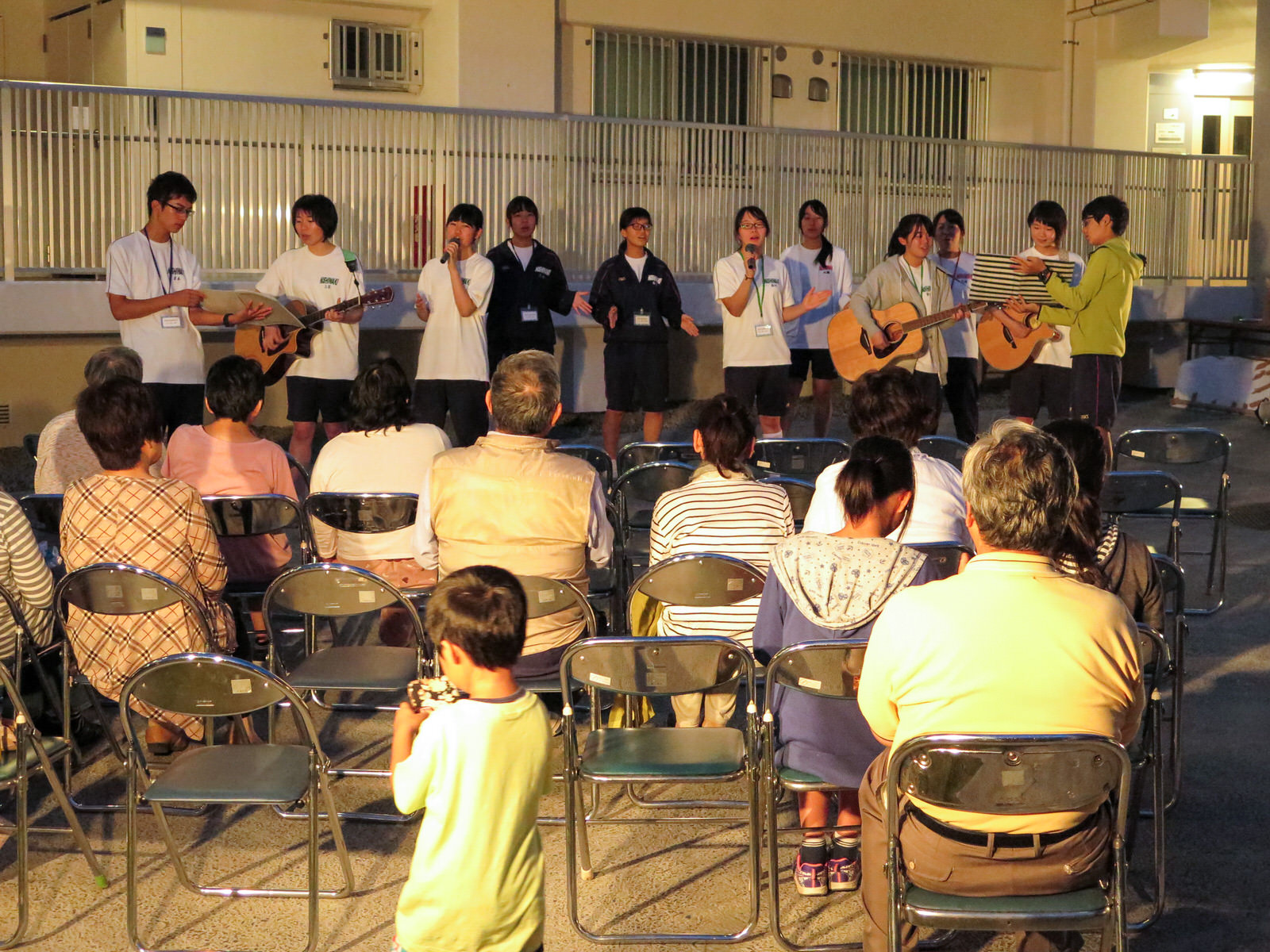 日が落ちてライトアップされた中庭で、観客の前に立った白いTシャツを着た高校生たちがギターを弾いたり、歌を歌っている様子の写真