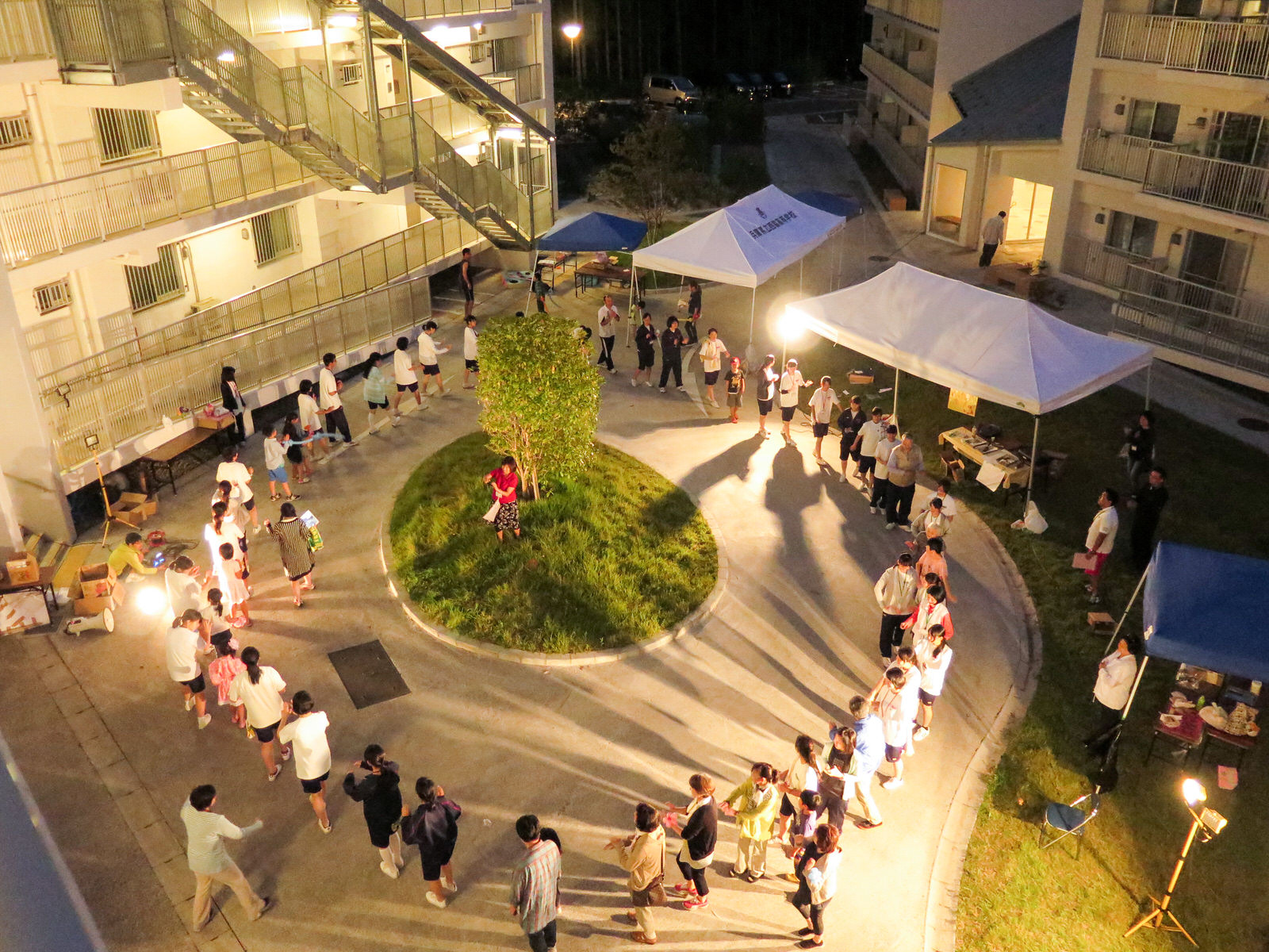 日が落ちてライトアップされた災害公営住宅の中庭で、丸い植え込みを囲んで輪になった高校生や地域の人々が盆踊りを楽しんでいる様子を上から写した写真