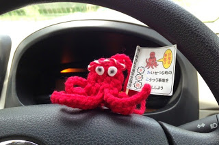 白い交通安全ののぼりを持った赤い毛糸で編まれたタコのぬいぐるみが車のハンドルの上に置かれている写真