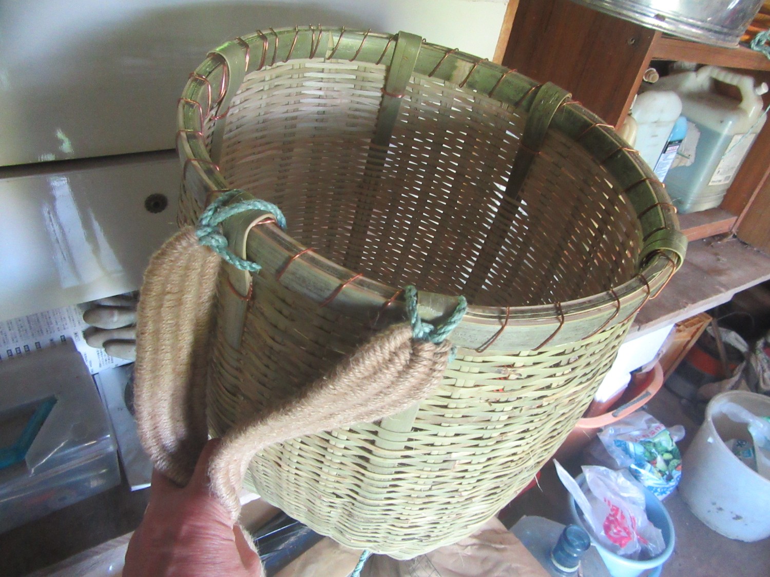 かごの上部には緑色の紐と柔らかそうな肩紐が取り付けられている、緑がかった竹で編まれた背負子を手に持っている写真