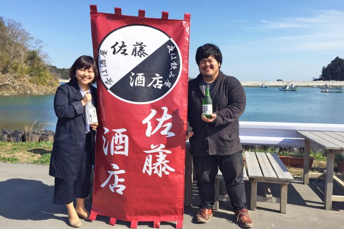 海を背景に「佐藤酒店」と書かれた赤い布看板をはさんで立った女性リポーターと佐藤さんがそれぞれ酒の瓶を持っている写真