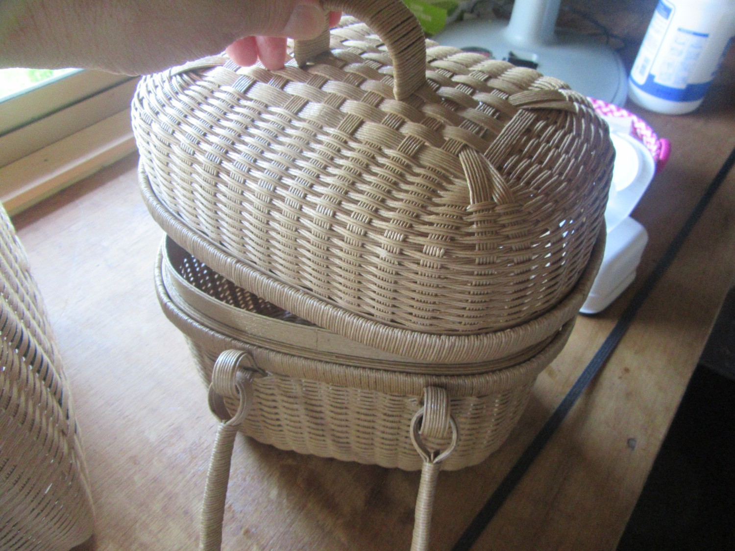 白っぽい素材でできた、丸みを帯びた形状で持ち手が付いた籠に乗せた、取っ手付きの蓋を手で持ち上げている写真
