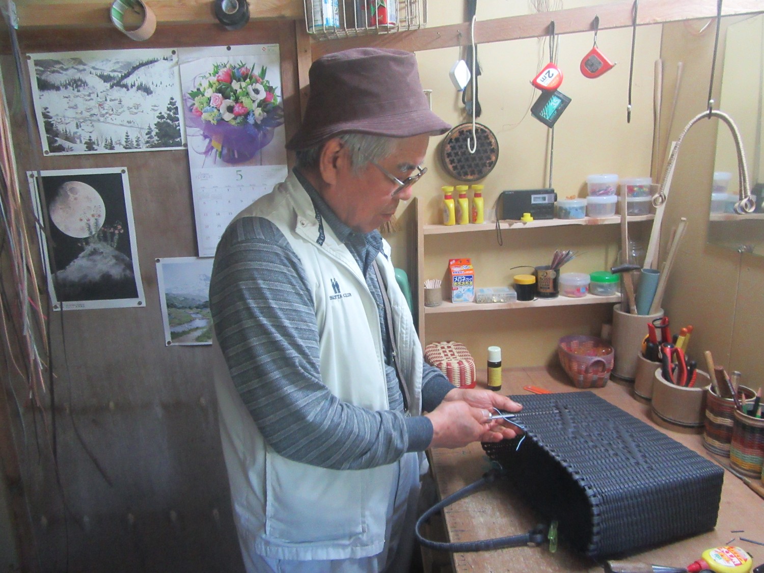 壁に絵や道具、材料などが並んでいる工房の作業台に立った佐藤さんが黒い竹かごを手作業で編んでいる様子の写真