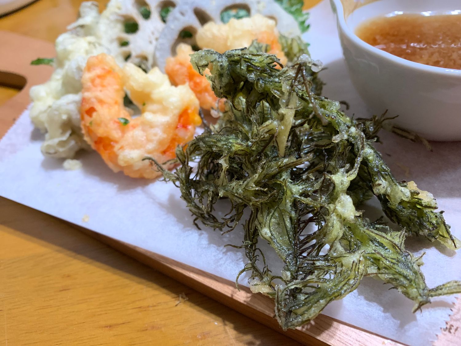 白い紙の上に並んだ天ぷらの中に、緑色の海藻を揚げたものがあり、えびや野菜の天ぷら、天つゆの小鉢も一緒に置かれている写真