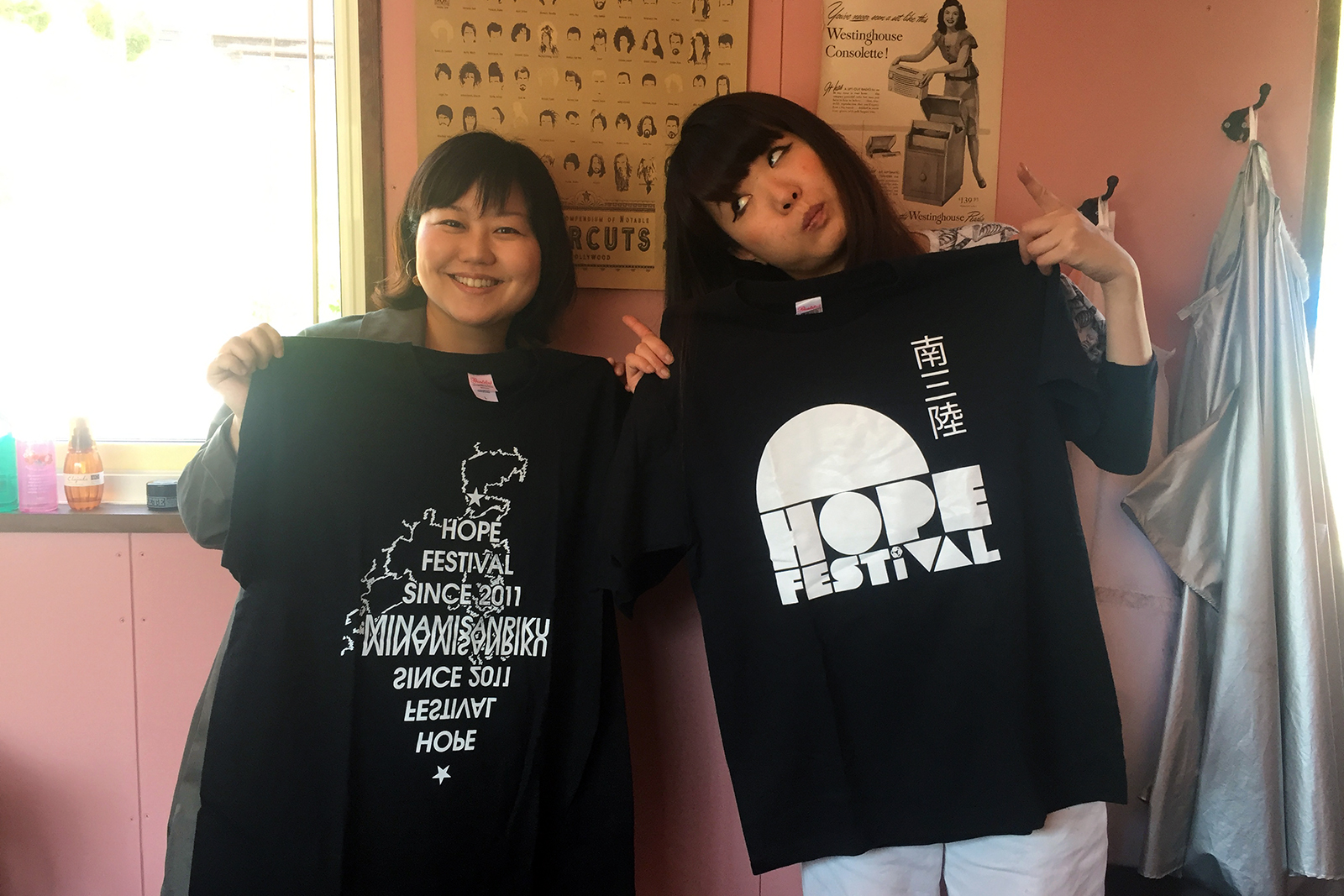 薄いピンク色の壁の前に並んで立った女性リポーターと伊藤さんが、黒い生地に白で「HOPE FESTIVAL」の文字などがデザインされたTシャツを広げて見せている写真