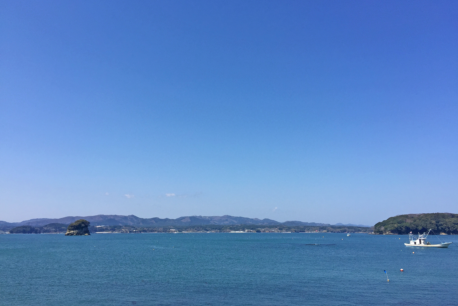 穏やかな青空の下、静かな海に小さな漁船が浮かび、遠くには緑の島々と山並みが広がる美しい海岸風景の写真