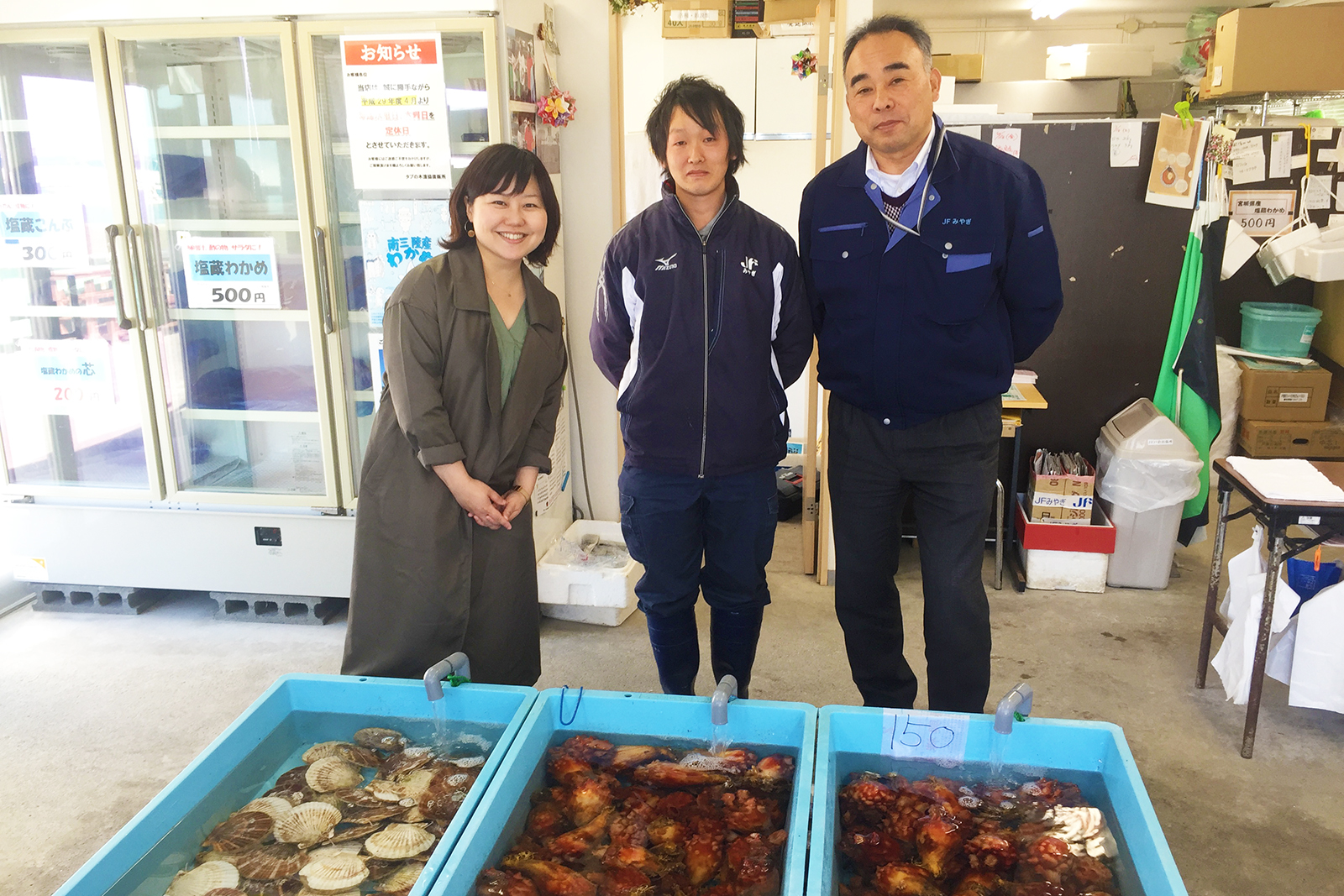 ホタテやホヤがそれぞれ入った3つの水色のいけすが並べられた後ろに女性リポーターと二人の男性が並んで立っている写真