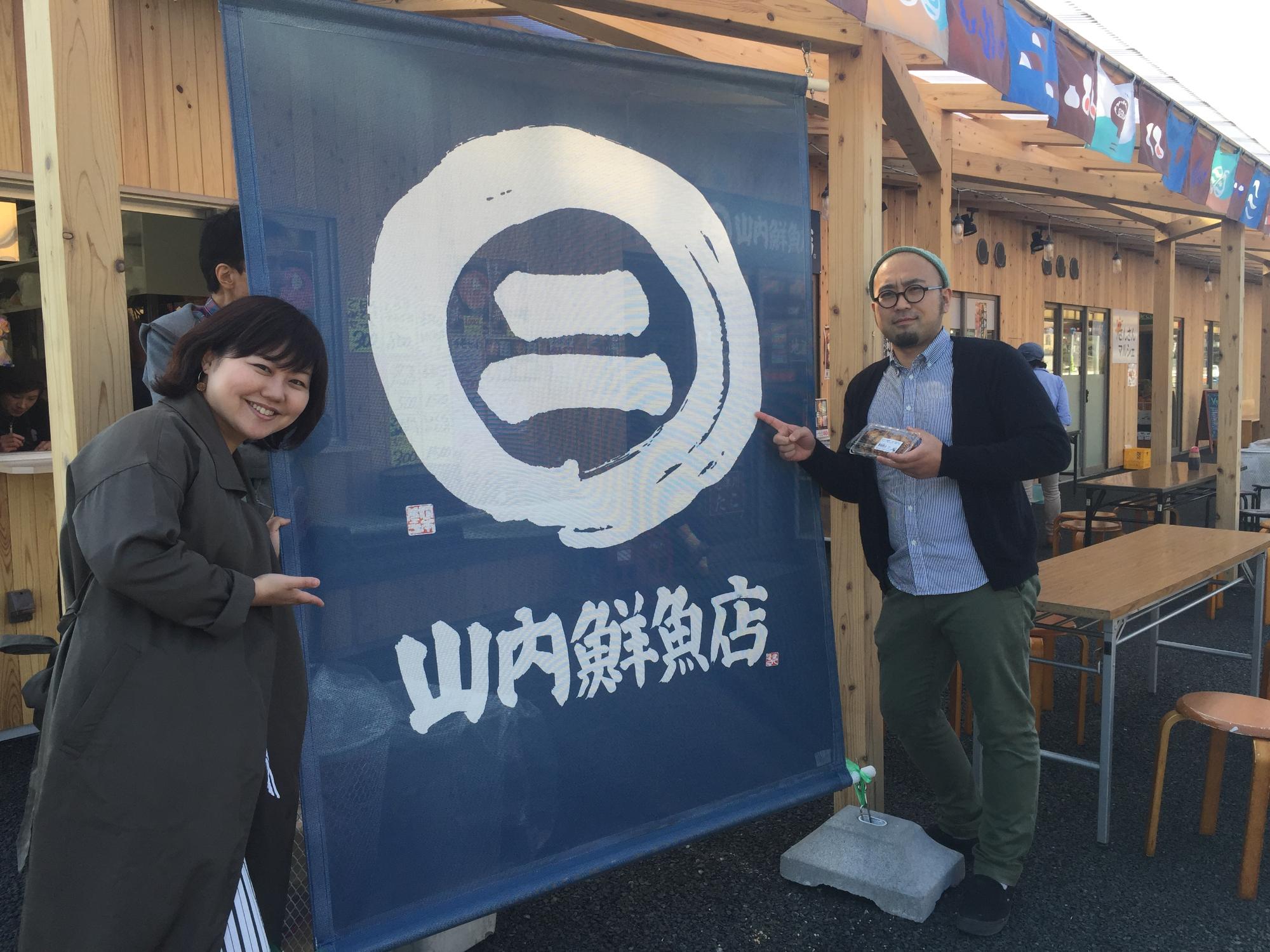 商店街の一角に設置された、紺色の生地に、白で書かれた丸の中に漢数字の2が書かれたロゴマークの下に「山内鮮魚店」と書かれた布製の看板の左右に女性リポーターと山内さんが立っている写真