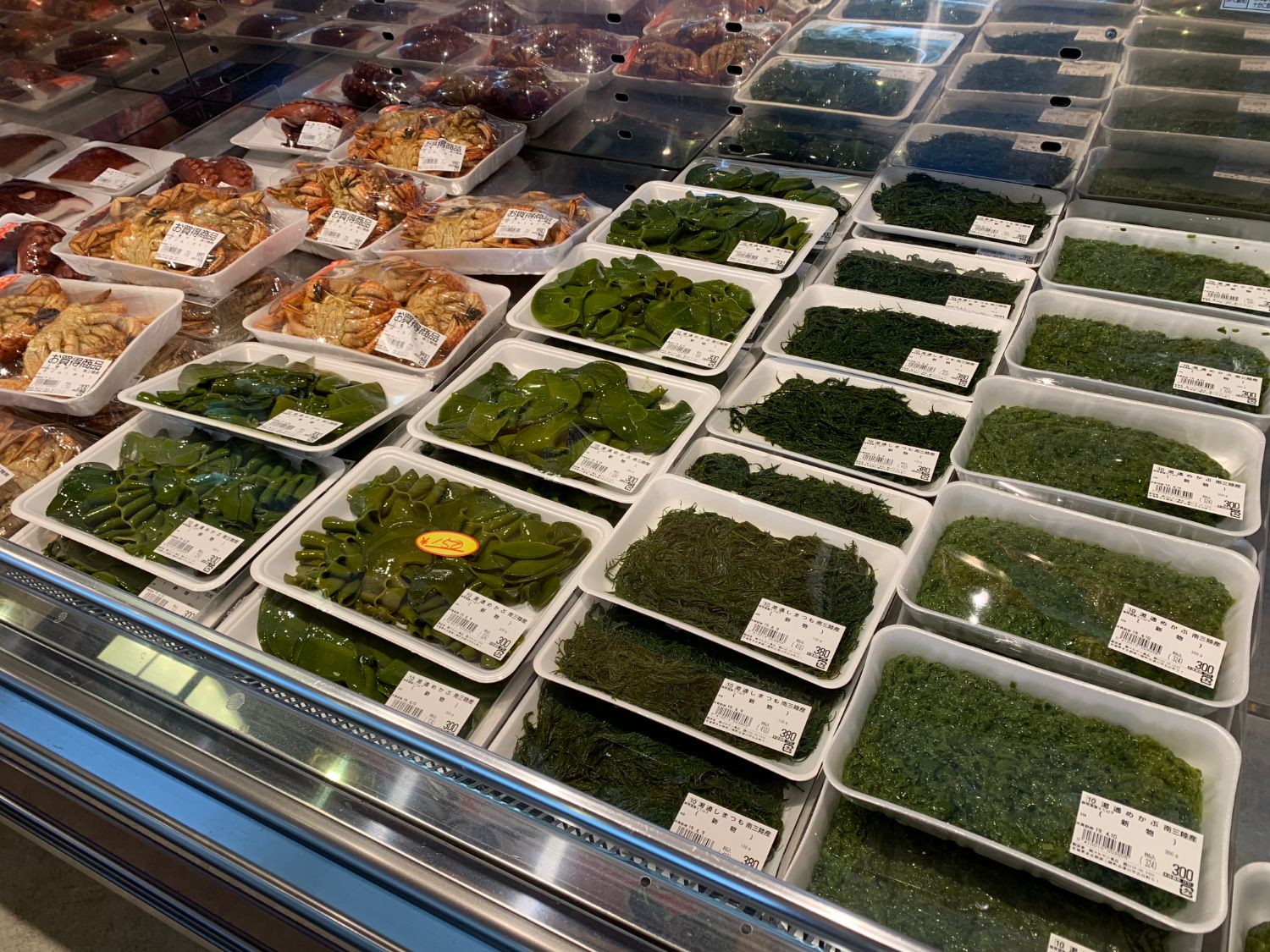 スーパーの冷蔵ケースに、値札やラベルが付いている様々な種類の緑色の海藻が白いトレーに小分けにされて並べられている写真