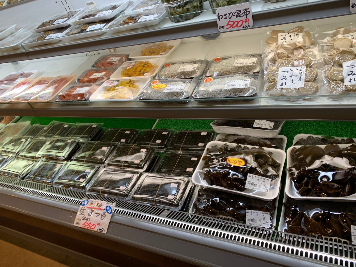 スーパーの冷蔵ケースに、佃煮や練り物、加工品などがパックに入れて並べられており、中央や手前には生まつもの製品がずらりと陳列されている写真