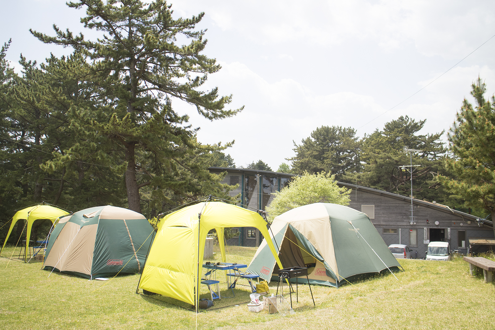 緑に囲まれた芝生のキャンプ場に、黄色いタープと緑色のテントが2つずつ設置されており、背後に木造の建物が見えている写真