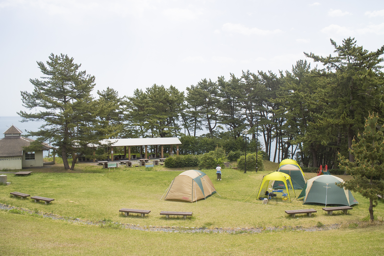 緑に囲まれたキャンプ場にテントやタープが設置され、屋根付きのスペースや小さい建物がある様子を上から撮影した写真