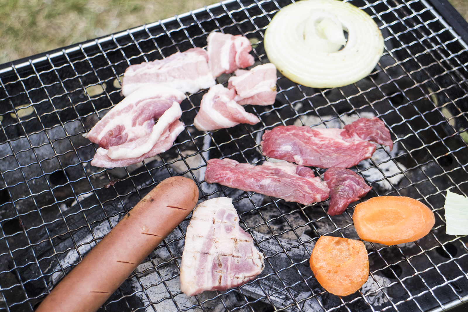 炭火焼きの網の上で肉とニンジン、玉ねぎ、ソーセージが焼かれている写真