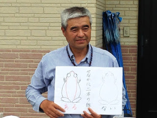 2匹のカエルの背中に二本の線があるバージョンと無いバージョンが描かれた白い紙を持ち説明している三塚先生の写真