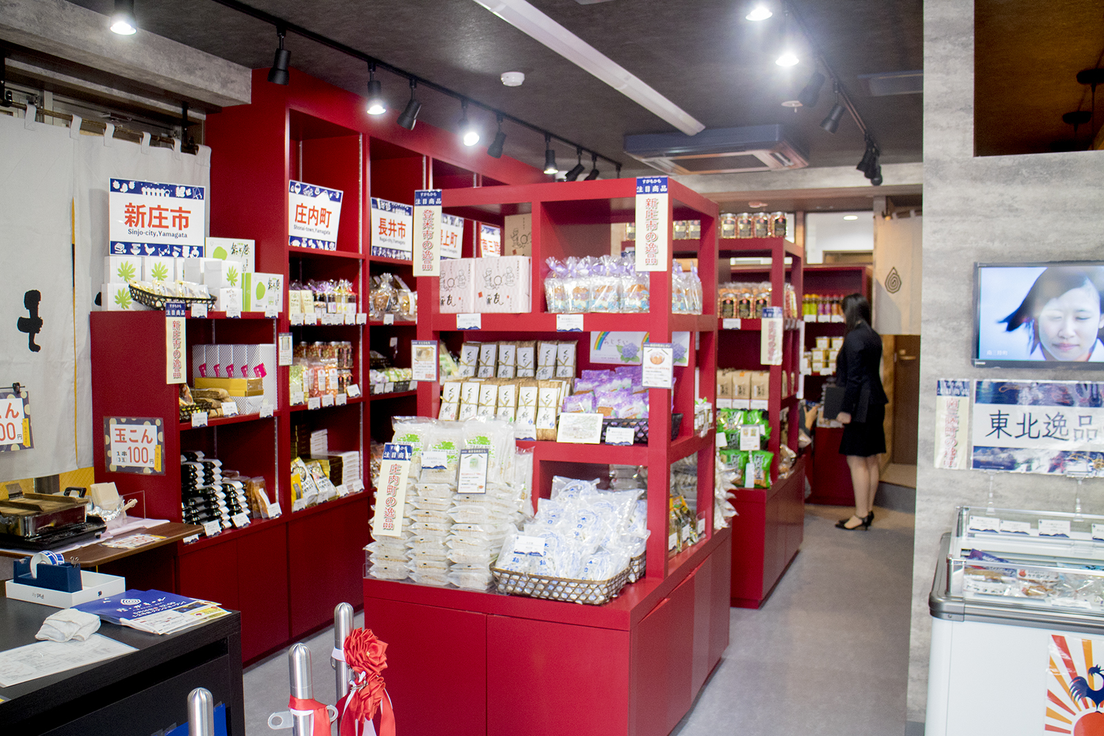 店内の赤い棚に地域特産品やお菓子が並び、奥で女性が商品を見ている様子の写真