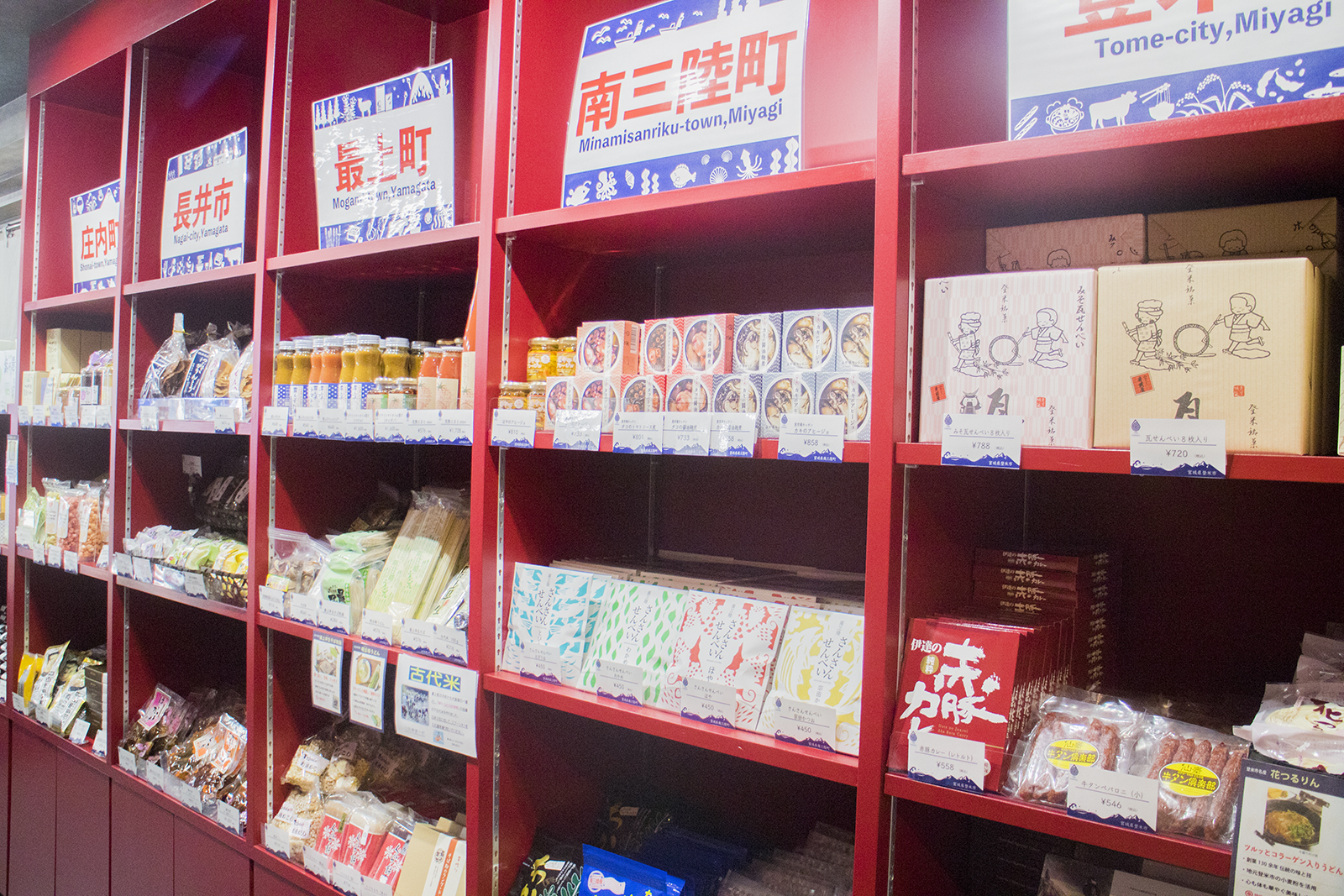 市や町の名前が掲げられた赤い棚に、それぞれの特産品が並べて陳列されている様子の写真